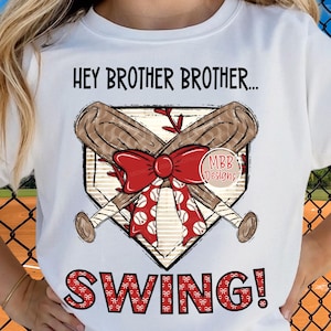 May include: White t-shirt with a baseball-themed graphic. The design features crossed baseball bats, a red bow, and the text "HEY BROTHER BROTHER...SWING!". The design includes a home plate shape and baseballs.