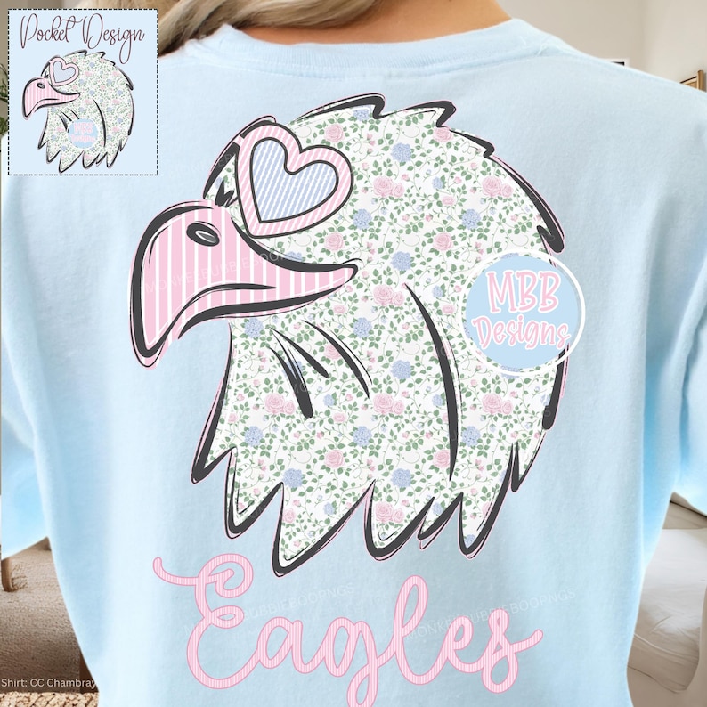 May include: Light blue shirt featuring a floral eagle design with pink and blue accents. The eagle has heart-shaped glasses and the word "Eagles" in pink script. A smaller version of the eagle is in the upper left corner.