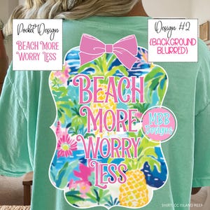May include: A light green t-shirt featuring a tropical design with the text "BEACH MORE WORRY LESS" in pink. The design includes a pink bow and tropical illustrations. The shirt also has a pocket design with the same text.