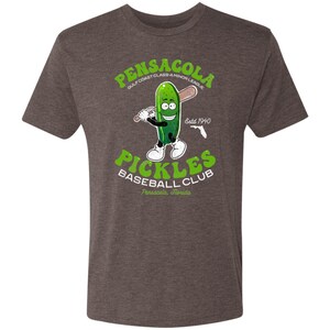 May include: A brown t-shirt featuring a green cartoon pickle holding a baseball bat. The text on the shirt reads "Pensacola Gulf Coast Class-A Minor League Estd 1940 Pickles Baseball Club Pensacola, Florida."