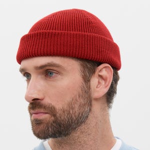 May include: A red ribbed knit beanie. The hat is a solid color and has a folded brim. The person in the image is wearing a light blue crew neck sweatshirt with a white undershirt.