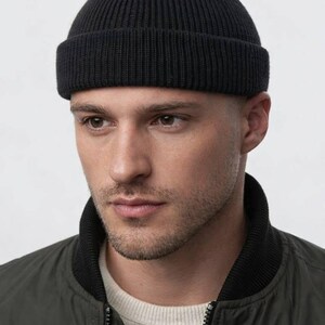 May include: A black ribbed knit beanie with a folded brim. The hat is worn by a person wearing a dark green bomber jacket with a black collar and a light-colored shirt underneath. The background is plain white.