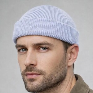 May include: A light blue ribbed knit beanie. The hat is worn by a person. The person is wearing a brown suede jacket over a cream-colored sweater. The background is a solid light gray.