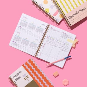 Quarterly Plans 90-Day Planner shown open to quarterly overview pages with the Tulip Wave cover design and planning accessories on a pink background.