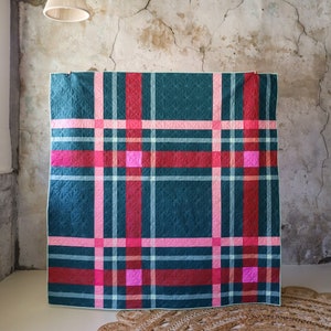 May include: A teal, pink, and red plaid quilt with a white background. The quilt is hanging on a wall with a rough, textured surface.