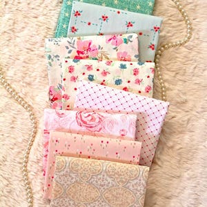 Puede incluir: Una pila de cuadrados de tela doblados con varios dise&ntilde;os florales y estampados. Los colores incluyen azul claro, rosa y crema. Las telas est&aacute;n dispuestas sobre una superficie esponjosa de color claro, con un collar de perlas drapeado a un lado.