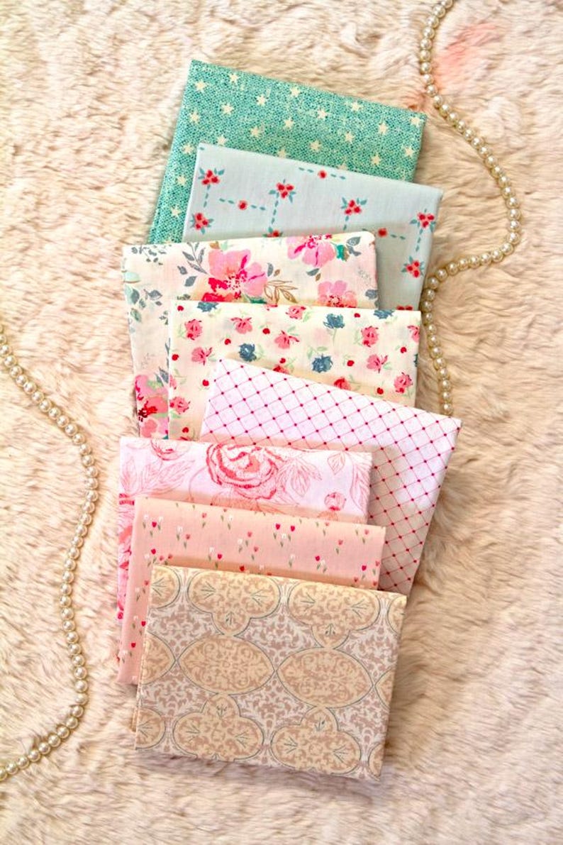 Puede incluir: Una pila de cuadrados de tela doblados con varios dise&ntilde;os florales y estampados. Los colores incluyen azul claro, rosa y crema. Las telas est&aacute;n dispuestas sobre una superficie esponjosa de color claro, con un collar de perlas drapeado a un lado.