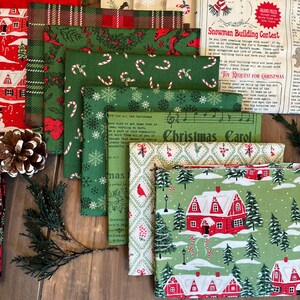 May include: A collection of folded fabric squares featuring various Christmas-themed patterns including red houses, green pine trees, candy canes, snowflakes, and text such as Snowman Building Contest and Christmas Carol, arranged in a layered stack.
