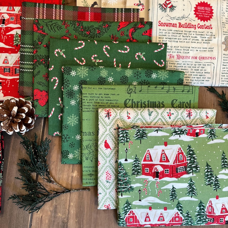 May include: A collection of folded fabric squares featuring various Christmas-themed patterns including red houses, green pine trees, candy canes, snowflakes, and text such as Snowman Building Contest and Christmas Carol, arranged in a layered stack.