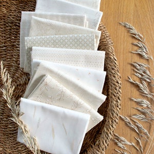 May include: A stack of eight white fabric squares with different subtle patterns in beige and brown. The fabric squares are arranged in a fan shape and are sitting in a woven basket.