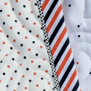 May include: Close-up of a quilted fabric with a white background. The fabric features a pattern of orange and navy blue polka dots and a striped section with orange, navy blue, and white stripes. A black and white dotted trim borders the striped section.