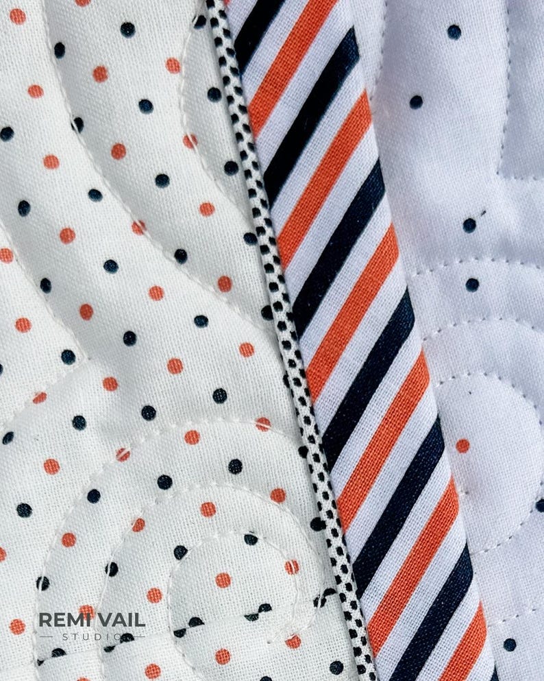 May include: Close-up of a quilted fabric with a white background. The fabric features a pattern of orange and navy blue polka dots and a striped section with orange, navy blue, and white stripes. A black and white dotted trim borders the striped section.