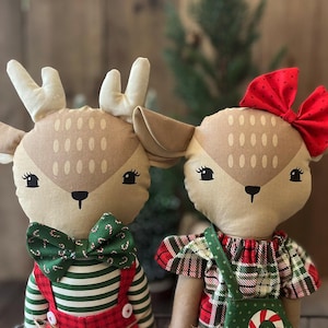 May include: Two plush deer dolls with tan fabric faces, embroidered eyes, and noses. The left deer wears a green candy cane print bowtie and red striped overalls. The right deer wears a red polka dot bow and a red and green plaid dress.