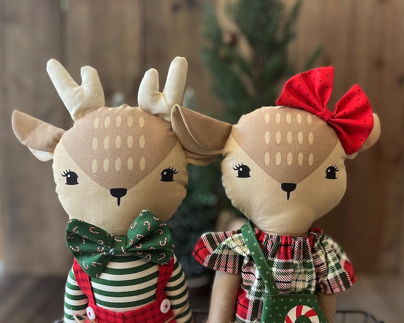 May include: Two plush deer dolls with tan fabric faces, embroidered eyes, and noses. The left deer wears a green candy cane print bowtie and red striped overalls. The right deer wears a red polka dot bow and a red and green plaid dress.