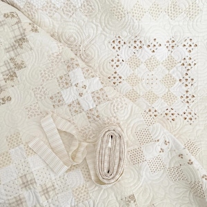 May include: A close-up of a white and cream quilt with a variety of patterns. The quilt is made of fabric squares with different floral prints. A roll of white fabric trim and a piece of white fabric ribbon are lying on top of the quilt.