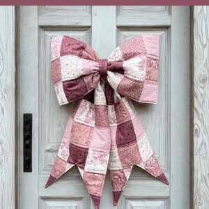 May include: A large, quilted bow in shades of pink and burgundy hangs on a white door. The bow is made of patchwork squares and has long, pointed tails. Text at the top reads "THE BIG GIANT QUILTED BOW". The text below says "Designed to be reversible, Scrappy and fat quarter friendly, 3 style options included".