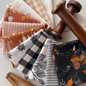 May include: A collection of folded fabric squares featuring various patterns including gingham, stripes, floral, and botanical prints in black, white, and rust orange, accompanied by two wooden spools and a wooden folding ruler.