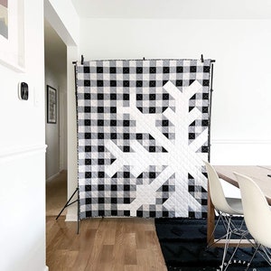 May include: A black and white gingham quilt with a tree design is displayed on a black stand. The quilt is made of square blocks and has a modern, geometric aesthetic. The room has hardwood floors and white walls.