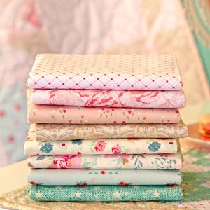 Puede incluir: Una pila de telas plegadas con varios dise&ntilde;os florales y estampados. Las telas son en tonos de rosa, crema y verde azulado. La tela superior tiene un patr&oacute;n de diamantes rojos. Las telas est&aacute;n apiladas sobre una superficie blanca.