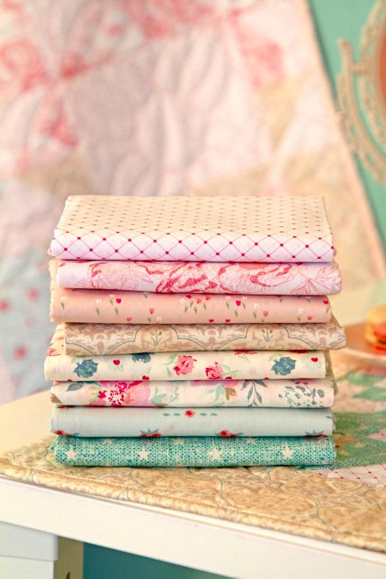 Puede incluir: Una pila de telas plegadas con varios dise&ntilde;os florales y estampados. Las telas son en tonos de rosa, crema y verde azulado. La tela superior tiene un patr&oacute;n de diamantes rojos. Las telas est&aacute;n apiladas sobre una superficie blanca.