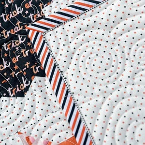 May include: Close-up of a Halloween-themed quilt with a black and white polka dot pattern and orange accents. The quilt features the words "trick or treat" in a swirling design. The border has orange and black stripes.