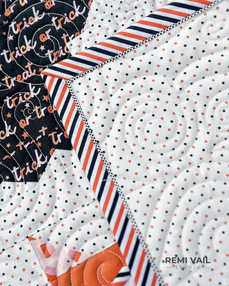 May include: Close-up of a Halloween-themed quilt with a black and white polka dot pattern and orange accents. The quilt features the words "trick or treat" in a swirling design. The border has orange and black stripes.