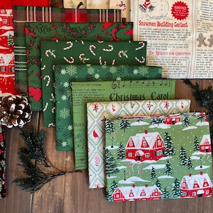 May include: A collection of folded cotton fabric squares featuring various holiday patterns including red and green plaid, candy canes, snowflakes, Christmas carols, and a winter village scene with red houses, pine trees, and snow-covered ground.