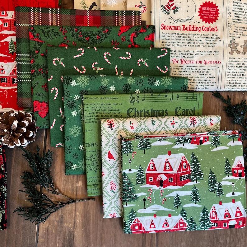 May include: A collection of folded cotton fabric squares featuring various holiday patterns including red and green plaid, candy canes, snowflakes, Christmas carols, and a winter village scene with red houses, pine trees, and snow-covered ground.