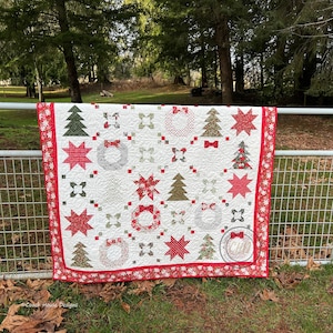 May include: A handmade Christmas quilt with a white background and a repeating pattern of red stars, wreaths, and Christmas trees. The quilt has a red border with a festive design and is displayed outdoors.