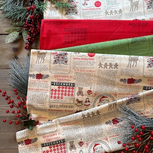 May include: A cream-colored fabric featuring a repeating Christmas-themed newspaper print with red and black text, illustrations of Santa, reindeer, gingerbread men, and a December calendar. The design includes sections titled Deck the Halls.