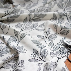 May include: White fabric with a black and white leaf pattern. The fabric is folded and a pair of black scissors is lying on top of it.