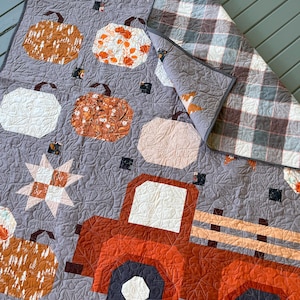 May include: A quilted blanket featuring a patchwork design with orange and white pumpkins, star shapes, and a large orange truck. The background is gray with a swirling stitch pattern, and the reverse side shows a gray and white plaid fabric.