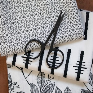 May include: Three pieces of white fabric with black geometric patterns. The top fabric has a repeating pattern of small black triangles. The middle fabric has a repeating pattern of black vertical lines and horizontal lines. The bottom fabric has a repeating pattern of black leaves. A pair of black fabric scissors is lying on top of the fabrics.