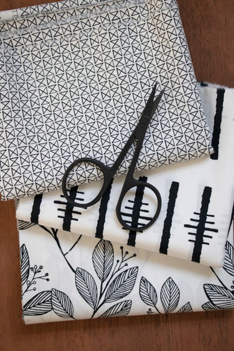 May include: Three pieces of white fabric with black geometric patterns. The top fabric has a repeating pattern of small black triangles. The middle fabric has a repeating pattern of black vertical lines and horizontal lines. The bottom fabric has a repeating pattern of black leaves. A pair of black fabric scissors is lying on top of the fabrics.