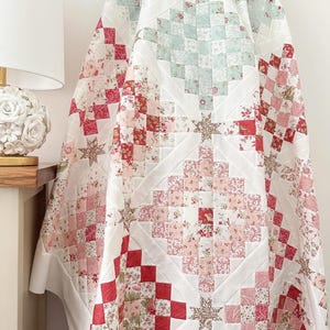 May include: A white quilt with a patchwork design featuring red, pink, and green floral patterns. The quilt is made up of squares and diamonds, and the fabric is soft and textured.