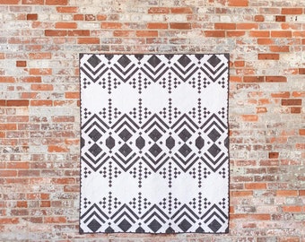 May include: A black and white geometric quilt with a diamond pattern hangs on a brick wall.