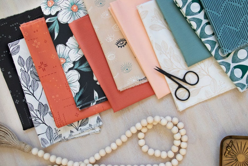 May include: A collection of fabric swatches featuring various patterns, including floral, geometric, and eye designs. The fabrics are in shades of black, white, green, blue, peach, and coral. A strand of wooden beads and a pair of black sewing scissors are also visible.