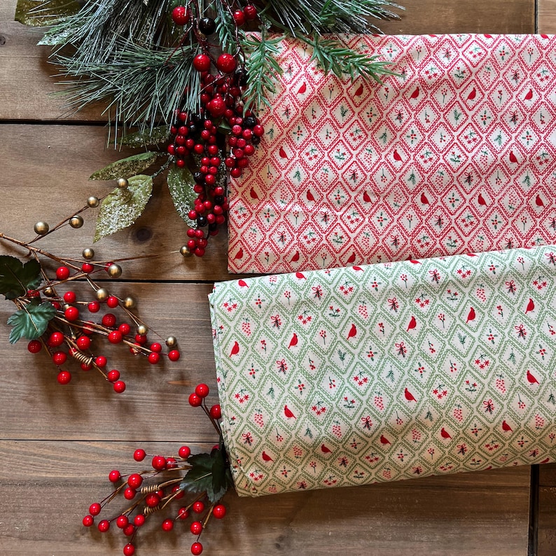 May include: Two folded pieces of fabric featuring a diamond grid pattern. One piece has a red background with white and green accents, while the other has a light green background with red and white accents. Both fabrics display small red birds.