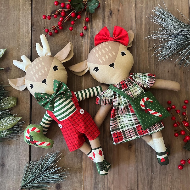 May include: Two plush deer dolls with tan faces and embroidered features. One deer wears a green and white striped shirt, red plaid overalls, and a candy cane bowtie. The other deer wears a red and green plaid dress with a red hair bow and a green bag.