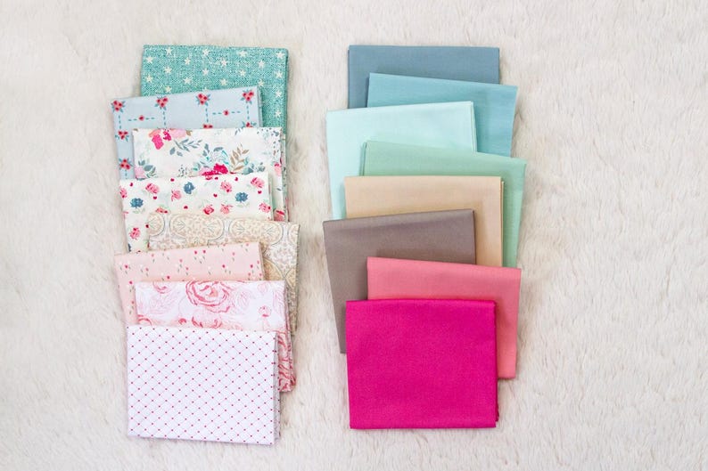 Puede incluir: Montones de cuadrados de tela en varios colores y patrones. La pila izquierda presenta dise&ntilde;os florales y de lunares en rosa, blanco y azul claro. La pila derecha incluye telas lisas en tonos de azul, rosa y beige.