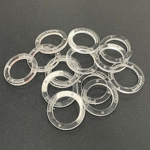 May include: A selection of transparent acrylic game tokens, each with engraved text. The circular tokens feature status effect words such as "Stunned", "Charmed", and "Poisoned". They are arranged on a dark background.