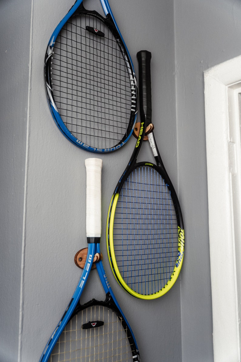 May include: Three tennis rackets hanging on a wall. The rackets are blue, black, and yellow. The rackets are hanging on wooden hooks.