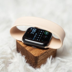 May include: A black Apple Watch with a light pink band is sitting on a wooden stand. The watch face shows the time as 7:00 and the date as "Tue, Mar 1".