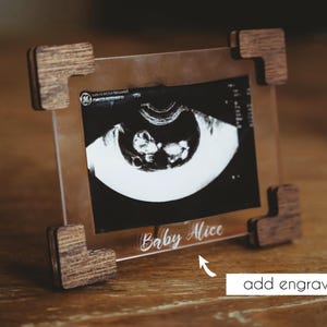 May include: A rectangular clear acrylic frame featuring dark brown wooden corner accents. The frame holds a black and white ultrasound image and displays the white engraved text Baby Alice at the bottom center of the clear acrylic surface.