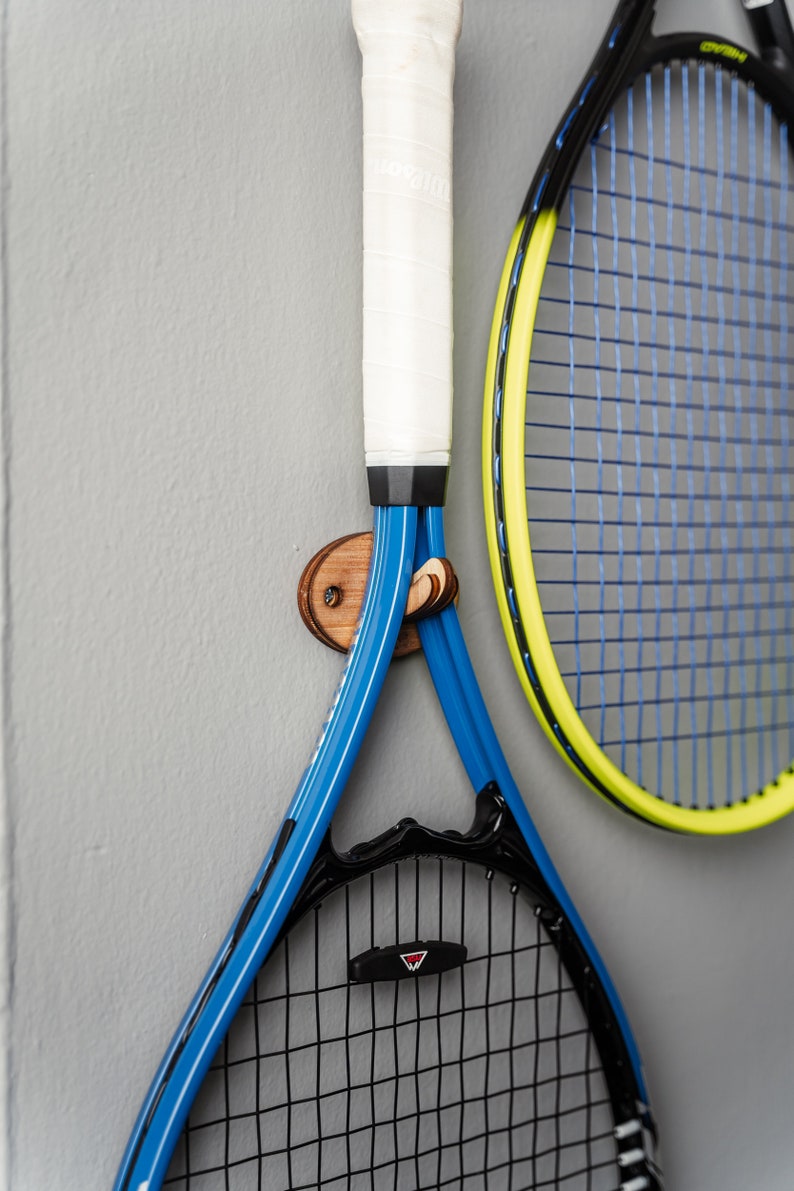 May include: Two tennis rackets hanging on a wooden wall hook. The racket on the left is blue and black with a white grip. The racket on the right is yellow and black with a blue and black string pattern.