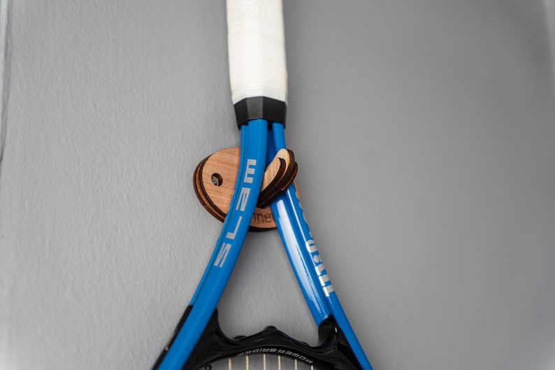 May include: Two blue tennis rackets with white lettering are hanging on a wooden wall mount. The mount is shaped like an oval and has a small hole in the center.