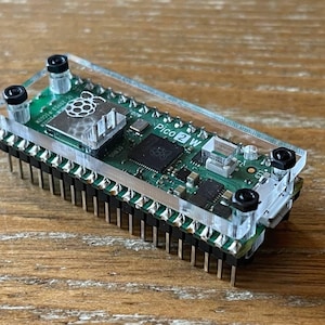 May include: A small, green circuit board labeled "Pico 2 W" with a Raspberry Pi logo. The board is mounted on a base with numerous pins and is encased in a clear acrylic cover, secured by black screws. The components are arranged on a wooden surface.