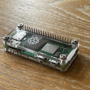 May include: A clear plastic case with a Raspberry Pi Zero W inside. The case has four silver screws and a Raspberry Pi logo on the board.