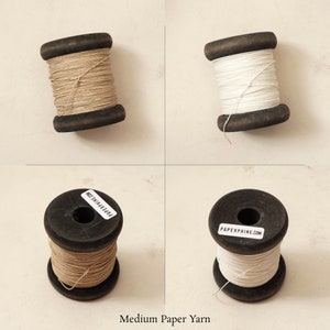 Puede incluir: Dos carretes de hilo de papel, uno de color marr&oacute;n natural y otro blanco, sobre un fondo beige claro. Los carretes son de madera y llevan una etiqueta de papel que dice "PAPERPHINE.COM".