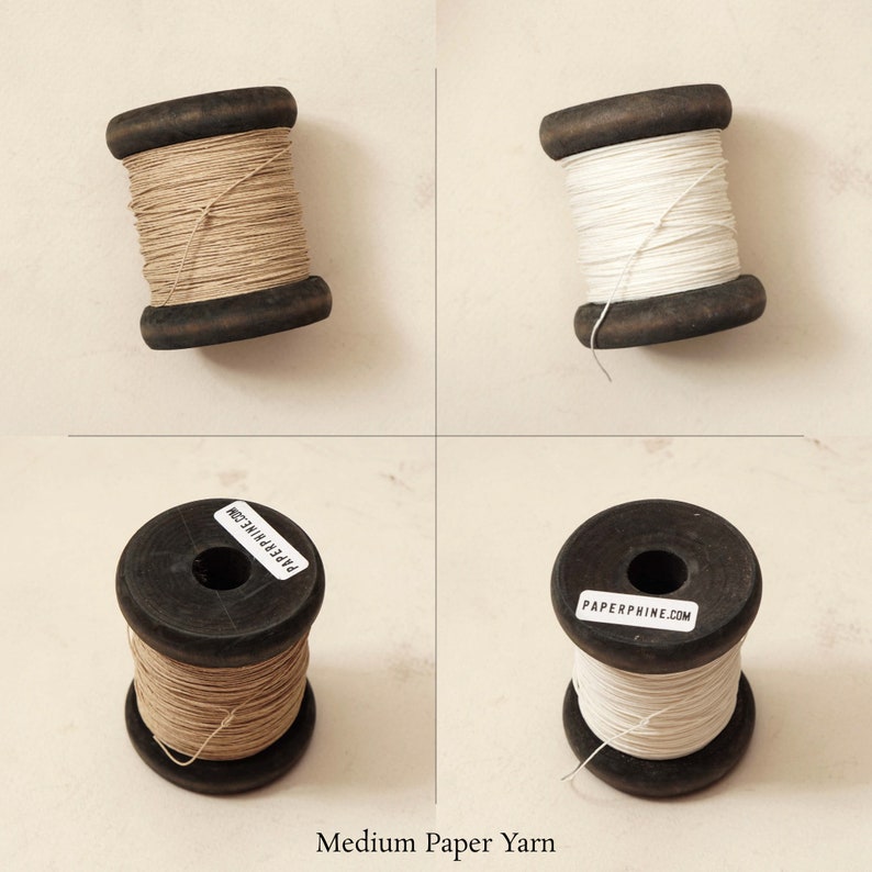 Puede incluir: Dos carretes de hilo de papel, uno de color marr&oacute;n natural y otro blanco, sobre un fondo beige claro. Los carretes son de madera y llevan una etiqueta de papel que dice "PAPERPHINE.COM".